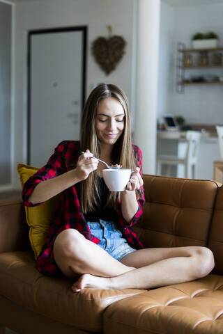 Entspannte junge Frau isst Müsli im Wohnzimmer zu Hause, lizenzfreies Stockfoto