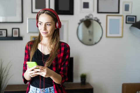 Junge Frau mit Mobiltelefon und Kopfhörern zu Hause, lizenzfreies Stockfoto