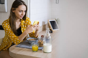 Junge Frau benutzt ein Handy und frühstückt in der Küche zu Hause - GIOF06702