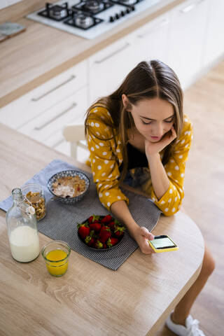 Junge Frau benutzt ein Handy und frühstückt in der Küche zu Hause, lizenzfreies Stockfoto