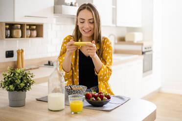 Glückliche junge Frau, die ein Handy benutzt und zu Hause in der Küche frühstückt - GIOF06706