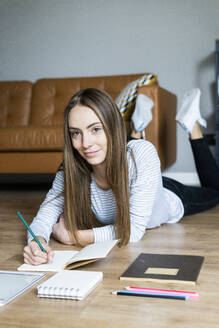 Portrait of smiling young woman lying on the floor at home taking notes - GIOF06713