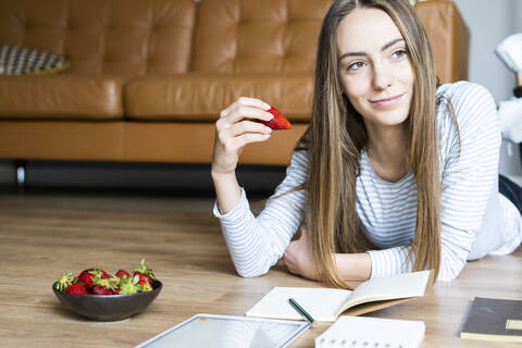 Relaxed young woman lying on the floor at home eating strawberries stock photo