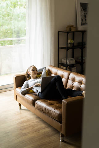 Junge Frau liegt zu Hause auf der Couch und benutzt einen Laptop, lizenzfreies Stockfoto