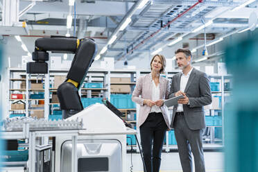Geschäftsmann und Geschäftsfrau in einer modernen Fabrikhalle mit Blick auf einen Roboter - DIGF07208