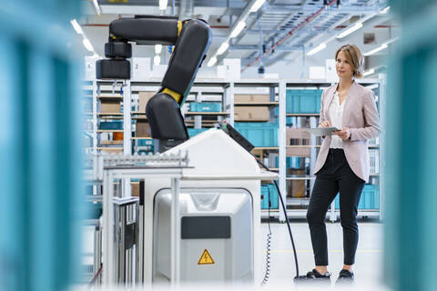 Geschäftsfrau mit Tablet in einer modernen Fabrikhalle mit Blick auf einen Roboter, lizenzfreies Stockfoto