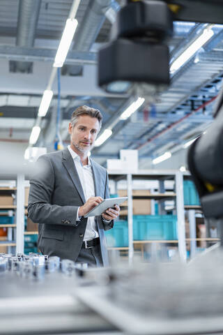 Geschäftsmann mit Tablet in einer modernen Fabrikhalle mit Blick auf einen Roboter, lizenzfreies Stockfoto