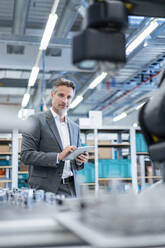 Geschäftsmann mit Tablet in einer modernen Fabrikhalle mit Blick auf einen Roboter - DIGF07212