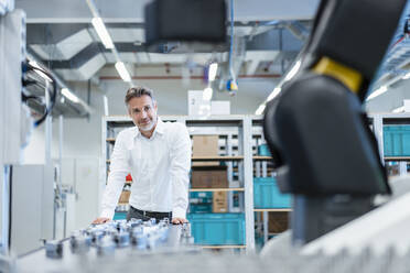 Confident businessman in a modern factory hall looking at robot - DIGF07215