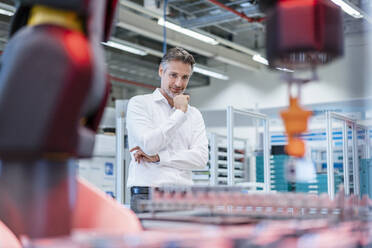 Confident businessman in a modern factory hall looking at robot - DIGF07221