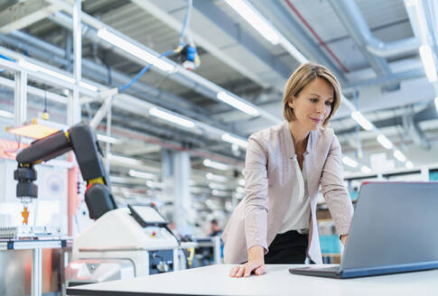 Businesswoman in a modern factory hall using laptop - DIGF07345