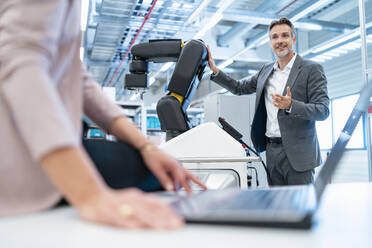 Geschäftsmann und Geschäftsfrau mit Laptop und Roboter in einer modernen Fabrikhalle - DIGF07351