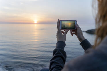 Spanien, Costa Brava, Nahaufnahme einer Frau, die mit ihrem Smartphone das Meer bei Sonnenaufgang fotografiert - AFVF03574