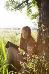 Porträt eines lächelnden Mädchens mit Buch, das sich auf einer Wiese an einen Baumstamm lehnt - LVF08162