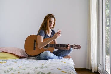 Junge Frau zu Hause, die mit Gitarre im Schlafzimmer chillt und ihr Smartphone benutzt - AFVF03592