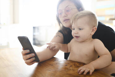 Portrait of little boy sitting with his mother at table looking at mobile phone - EYAF00288