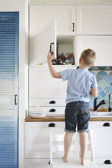 Rückenansicht eines kleinen Jungen, der auf einem Stuhl in der Küche steht und in einen Schrank schaut - EYAF00289