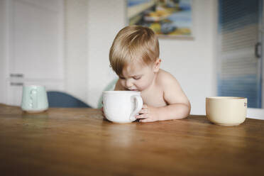 Shirtless little boy sitting at kitchen table looking into cup - EYAF00305