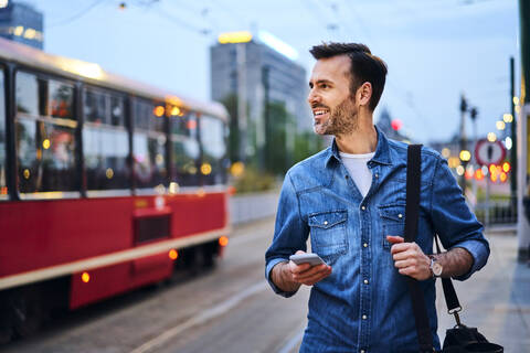 Mann benutzt sein Smartphone, während er abends auf eine Straßenbahn wartet, lizenzfreies Stockfoto