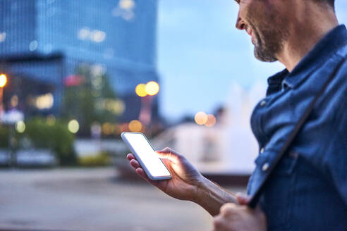 Nahaufnahme eines Mannes, der sein Smartphone benutzt, während er abends in der Stadt unterwegs ist - BSZF01086