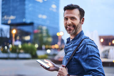 Portrait of smiling man standing in the city during evening and using smartphone - BSZF01087
