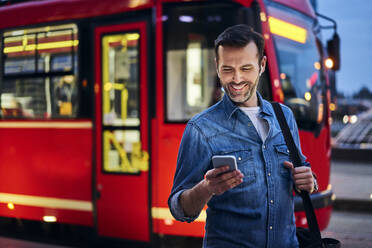 Smiling man using smartphone in the city with tram riding in background - BSZF01088