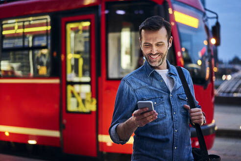 Lächelnder Mann mit Smartphone in der Stadt mit Straßenbahn im Hintergrund - BSZF01088