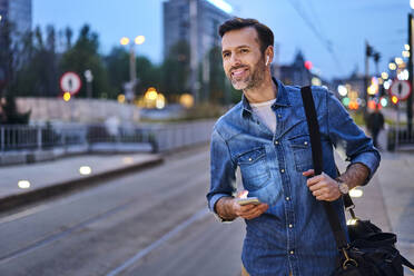 Lächelnder Mann mit drahtlosen Kopfhörern und Smartphone, der an der Straßenbahnhaltestelle wartet, während er abends nach der Arbeit pendelt - BSZF01090