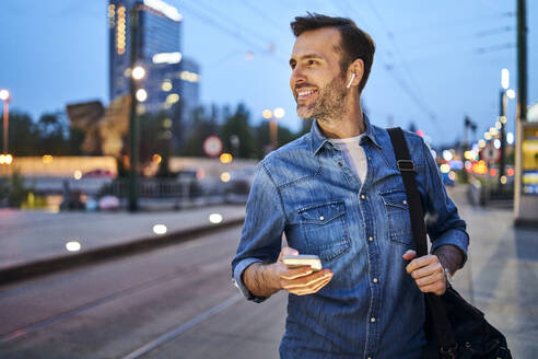 Lächelnder Mann, der sein Smartphone benutzt und über drahtlose Kopfhörer Musik hört, während er an der Straßenbahnhaltestelle auf den Feierabend wartet - BSZF01091