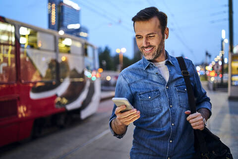 Man using smartphone and listening to music while standing at tram stop in the evening stock photo