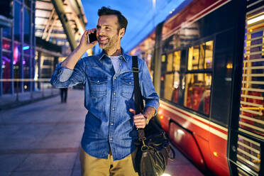 Mann telefoniert abends in der Stadt mit Straßenbahnfahrt im Hintergrund - BSZF01094