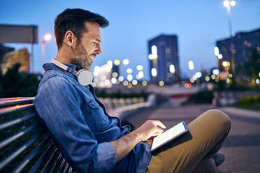Mann benutzt ein Tablet, während er abends auf einer Bank in der Stadt sitzt - BSZF01096