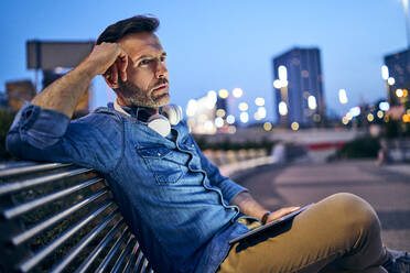 Worried man looking away while sitting on public bench with tablet - BSZF01097