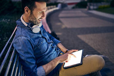 Man using tablet while sitting on a bench in the city in the evening - BSZF01098
