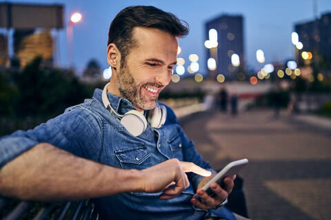 Lächelnder Mann, der sein Smartphone benutzt, während er abends auf einer Bank sitzt, lizenzfreies Stockfoto