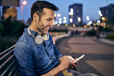 Smiling man using his smartphone while sitting on a bench in the evening - BSZF01101