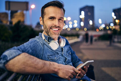 Porträt eines lächelnden Mannes, der sein Smartphone benutzt, während er abends auf einer Bank sitzt, lizenzfreies Stockfoto