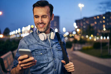 Smiling man using his smartphone in the city in the evening - BSZF01103