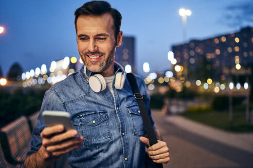 Lächelnder Mann, der sein Smartphone in der Stadt am Abend benutzt - BSZF01103