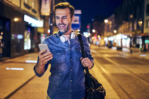 Lächelnder Mann benutzt sein Smartphone in der nächtlichen Stadt, lizenzfreies Stockfoto