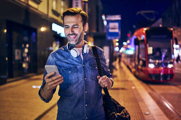 Smiling man using his smartphone in the city at night - BSZF01105
