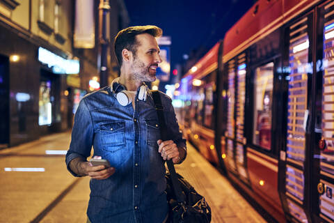 Lächelnder Mann, der nachts in der Stadt sein Smartphone benutzt, während er auf die Straßenbahn wartet, lizenzfreies Stockfoto
