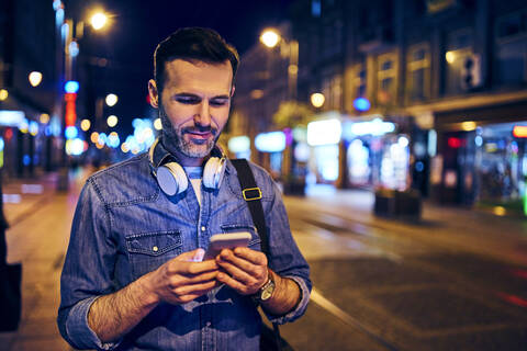 Lächelnder Mann, der nachts in der Stadt sein Smartphone benutzt, während er auf die Straßenbahn wartet, lizenzfreies Stockfoto