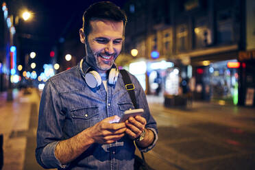 Smiling man using his smartphone in the city at night while waiting for the tram - BSZF01108