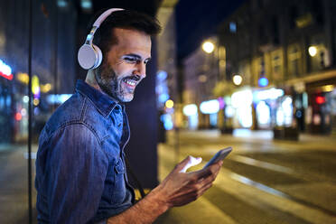 Smiling man with headphones using smartphone while waiting for night bus in the city - BSZF01110