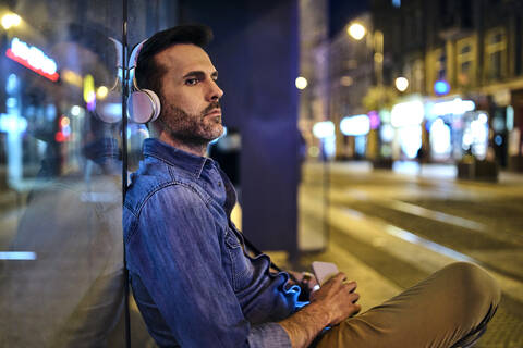 Ernster Mann mit drahtlosen Kopfhörern, der in der Stadt auf den Nachtbus wartet, lizenzfreies Stockfoto