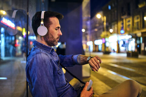 Seriöser Mann mit drahtlosen Kopfhörern, der in der Stadt auf den Nachtbus wartet und die Zeit überprüft, lizenzfreies Stockfoto