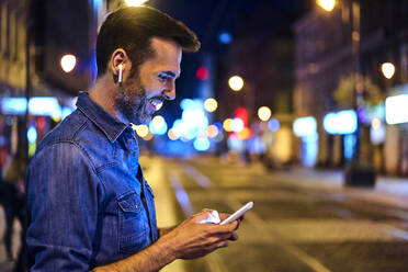 Man with wireless headphones using smartphone in the city at night - BSZF01115