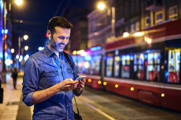 Lächelnder Mann mit drahtlosen Kopfhörern, der sein Smartphone benutzt, während er nachts auf die Straßenbahn wartet - BSZF01116