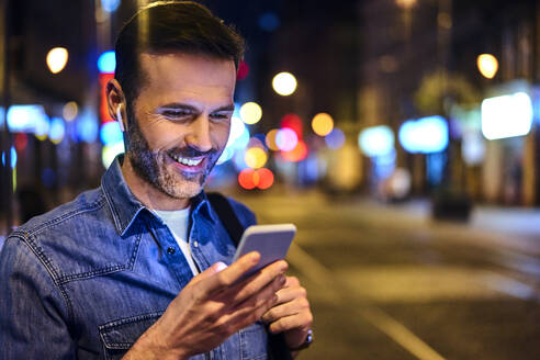 Lächelnder Mann mit drahtlosen Kopfhörern und Smartphone in der nächtlichen Stadt - BSZF01117